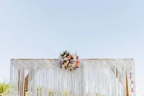 Arco de ceremonia con tejido de macramé blanco y flores en tonos pastel bajo el cielo despejado.