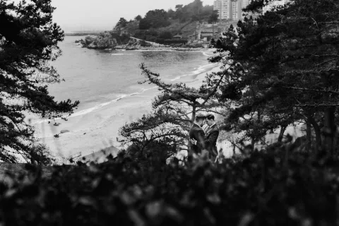 Vista en blanco y negro de una bahía enmarcada por árboles, con dos personas con traje juntas en el mirador.