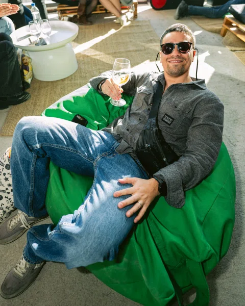 Hombre con gafas de sol sentado en un puff verde y sosteniendo una copa.