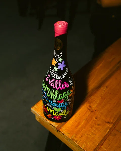 Botella decorada con flores y letras de colores sobre un banco de madera.