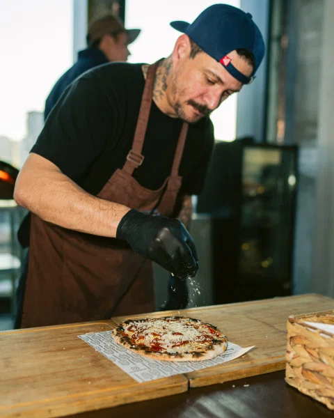 Hombre con delantal y guantes espolvorea un ingrediente sobre una pizza.