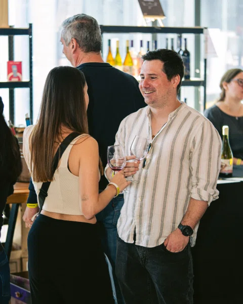 Dos personas conversan con copas de vino junto a una estantería de botellas.