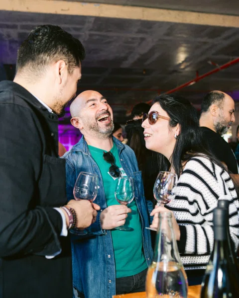 Tres personas conversan y ríen mientras sostienen copas de vino.