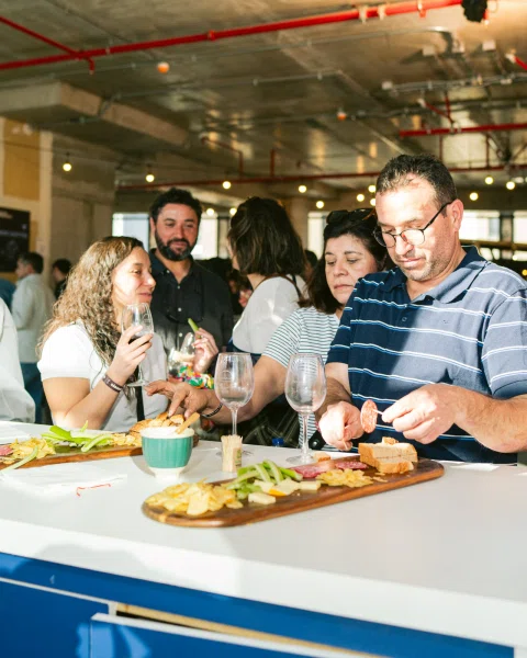 Personas junto a una barra con tablas de aperitivos, embutidos y copas.