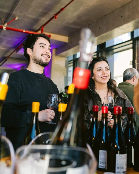 Dos personas sonríen mientras sostienen copas entre botellas en primer plano.