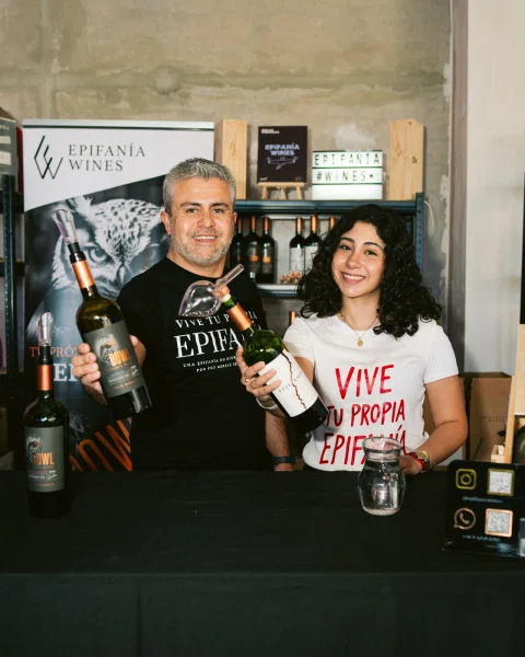 Dos personas sonríen detrás de una mesa con botellas de vino.
