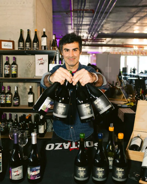Hombre sostiene tres botellas hacia la cámara detrás de una mesa con botellas.