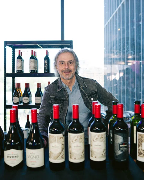 Hombre sentado detrás de una mesa con botellas de vino y una estantería al fondo.