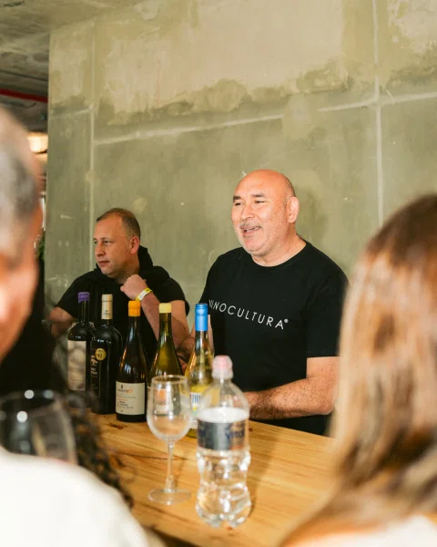 Hombre detrás de una mesa con botellas de vino conversa con asistentes desenfocados en primer plano.