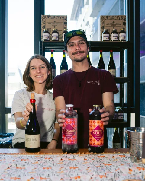 Dos personas sonríen detrás de una mesa con botellas, frente a una estantería con productos Aconcagua.