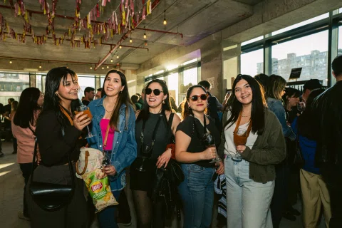 Cinco mujeres posan con copas entre asistentes y decoraciones colgantes.