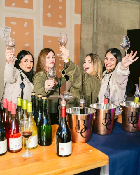 Cuatro mujeres levantan copas vacías detrás de una mesa con botellas y baldes metálicos.