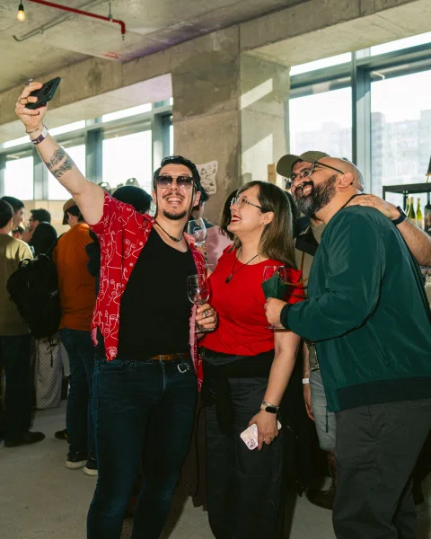 Grupo de personas sonríe para una selfie; varias sostienen copas.