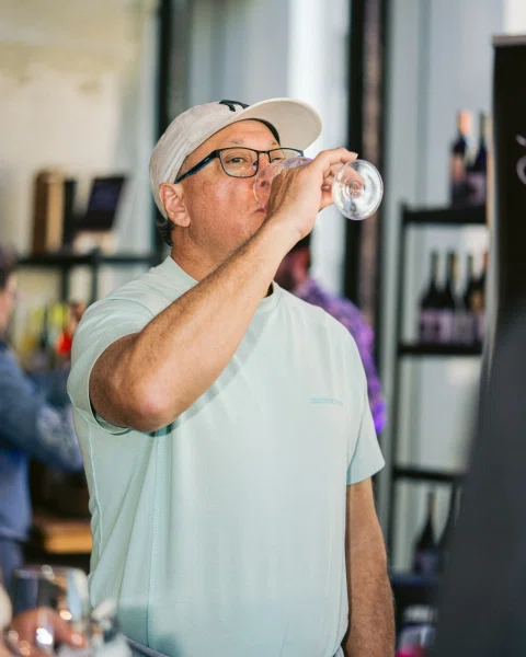 Hombre con gorra y lentes bebe de una copa frente a una estantería con botellas.