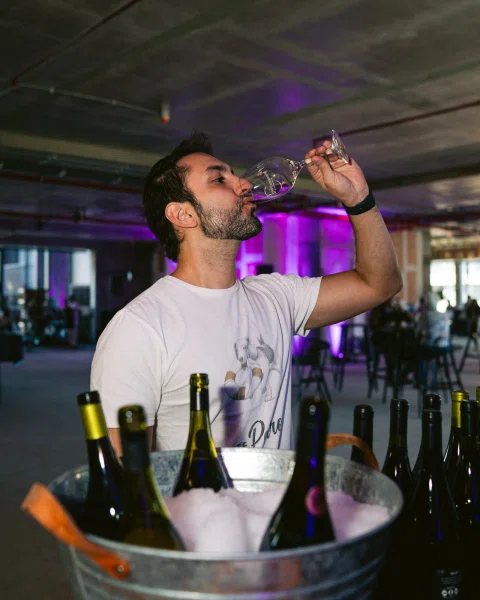 Hombre bebiendo de una copa frente a una cubeta con botellas y hielo.