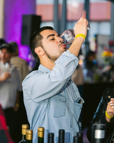Hombre con camisa de mezclilla bebe de una copa, con botellas desenfocadas en primer plano.
