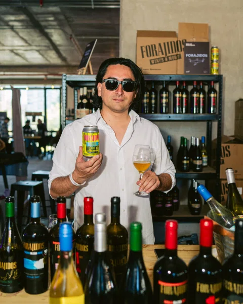 Hombre con gafas de sol sostiene una lata y una copa con bebida ámbar frente a botellas.