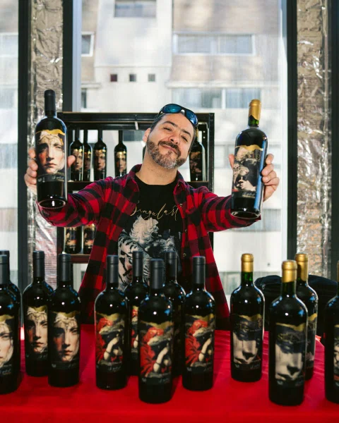 Hombre sostiene dos botellas de vino frente a una mesa cubierta de botellas.