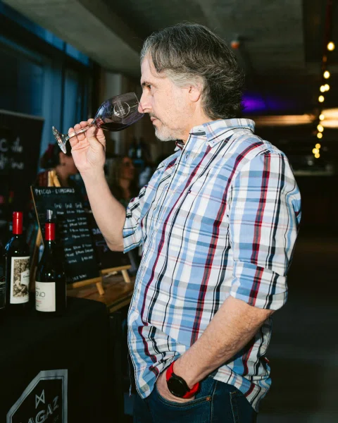 Hombre con camisa a cuadros oliendo una copa de vino junto a una mesa con botellas.