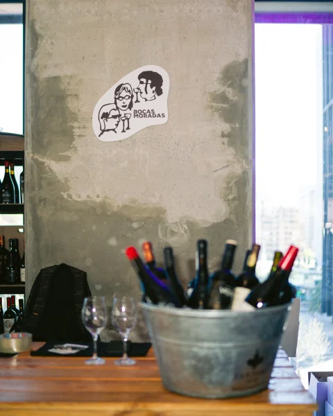 Letrero de Bocas Moradas sobre una mesa con copas y una cubeta llena de botellas.