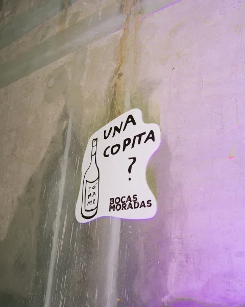 Letrero con el texto «Una copita?» y «Bocas Moradas» sobre una pared de concreto iluminada en violeta.