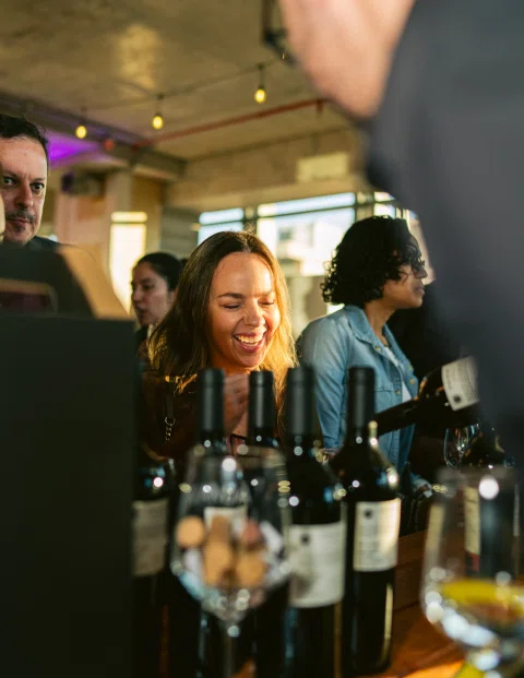 Mujer sonríe detrás de varias botellas de vino sobre una mesa.