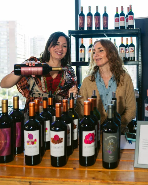 Dos mujeres sonríen mientras una sirve vino tinto en una copa, rodeadas de botellas.