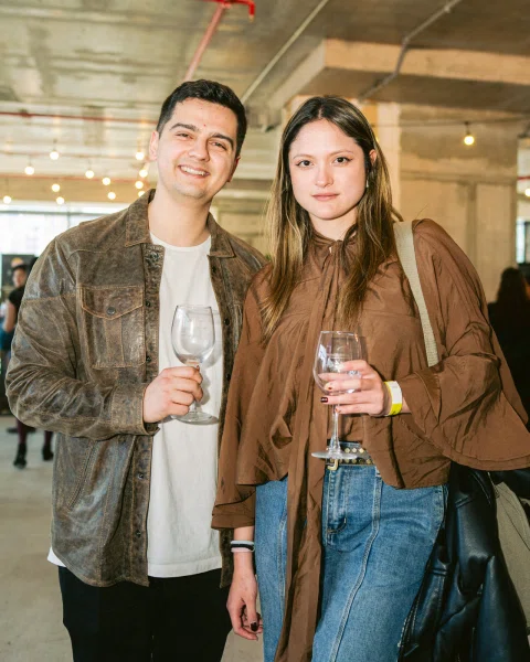 Dos personas posan con copas de vino bajo luces colgantes.