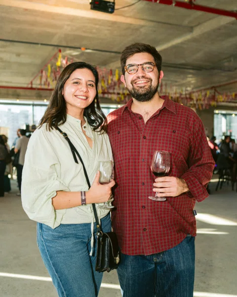 Dos personas posan sonrientes con copas de vino frente a decoración de cintas.