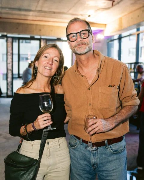 Dos personas posan sonrientes con copas de vino en un espacio interior.
