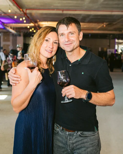 Dos personas posan abrazadas con copas de vino tinto.