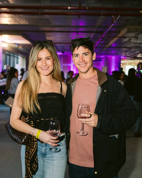Dos personas sonríen a la cámara mientras sostienen copas de vino en un espacio con iluminación violeta.