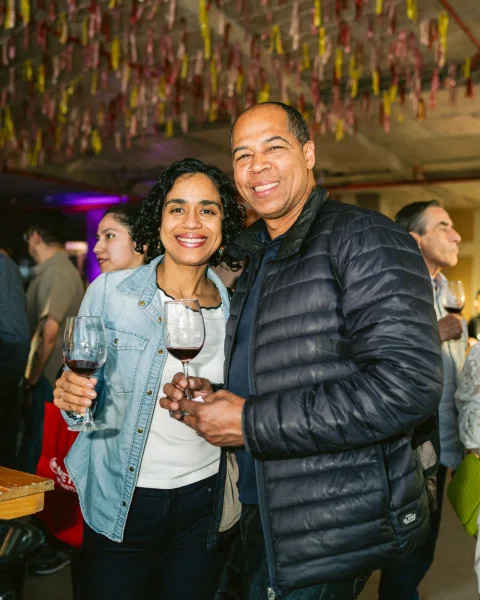 Dos personas sonríen a la cámara sosteniendo copas con vino tinto.