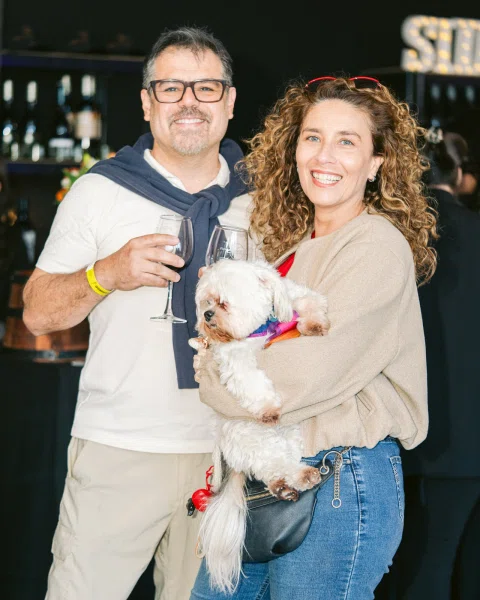 Dos personas sonríen con copas mientras una de ellas sostiene un perro pequeño.