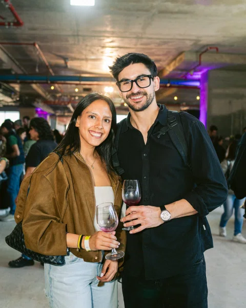 Dos personas posan sonrientes con copas de vino en un espacio interior concurrido.