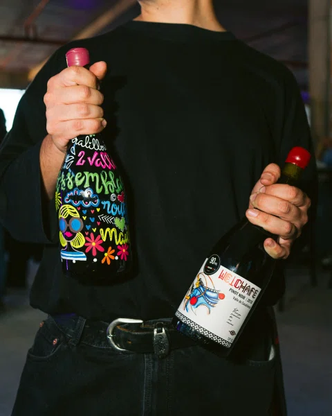Persona vestida de negro sosteniendo dos botellas de vino, una decorada y otra con etiqueta blanca.
