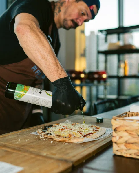 Hombre con guantes vierte aceite sobre una pizza en una tabla de madera.