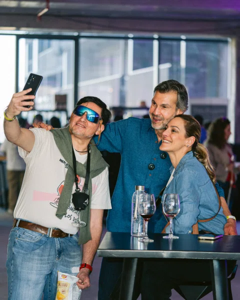 Tres personas sonríen para una selfie junto a una mesa alta con copas.
