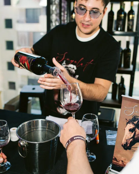 Persona sirviendo vino tinto en una copa sobre una mesa con botellas.