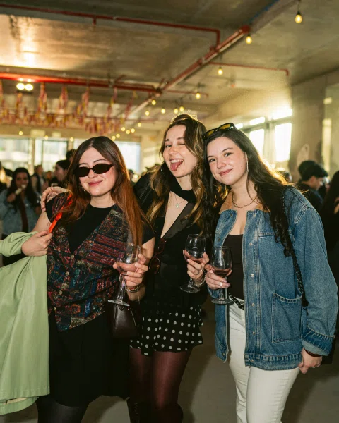 Tres mujeres posan sonrientes con copas en un espacio interior iluminado.