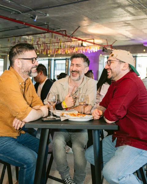 Tres hombres ríen y conversan alrededor de una mesa con copas y un plato de comida.