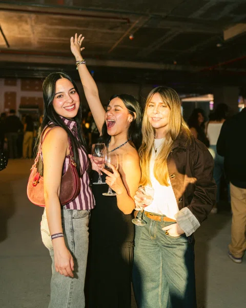Tres mujeres posan con copas mientras una levanta un brazo.