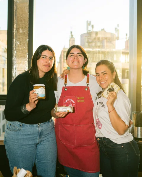 Tres mujeres posan mostrando un frasco y productos envueltos frente a una ventana.