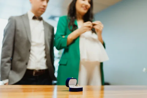 Caja azul abierta con dos anillos sobre una mesa, con un hombre y una mujer desenfocados al fondo.