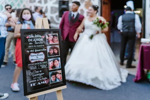 Cartel negro con fotografías y frases en primer plano, con un hombre de traje burdeos y una mujer de vestido blanco desenfocados al fondo.