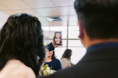 Celular sostenido en primer plano muestra el rostro de una mujer mientras dos personas aparecen desenfocadas detrás.