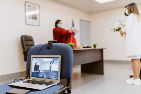 Computador portátil sobre una silla muestra una videollamada, con dos mujeres desenfocadas al fondo de la sala.