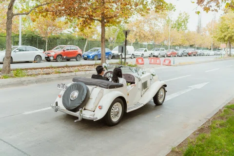 Automóvil convertible blanco con dos personas en su interior circula por una calle, visto desde atrás.