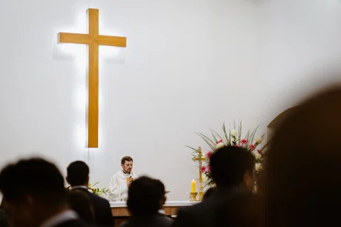 Gran cruz dorada sobre el altar de una iglesia, con un hombre de vestimenta litúrgica al fondo y dos personas vistas de espaldas en primer plano.