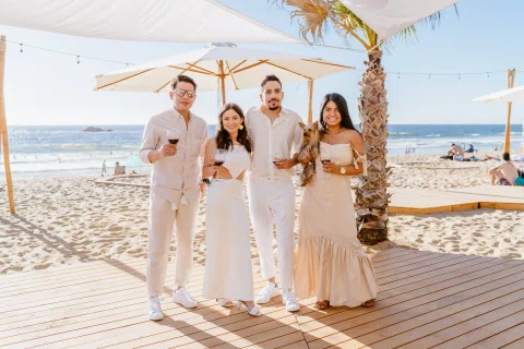 Cuatro personas posan en una terraza junto a la playa; una de ellas sostiene un perro pequeño.